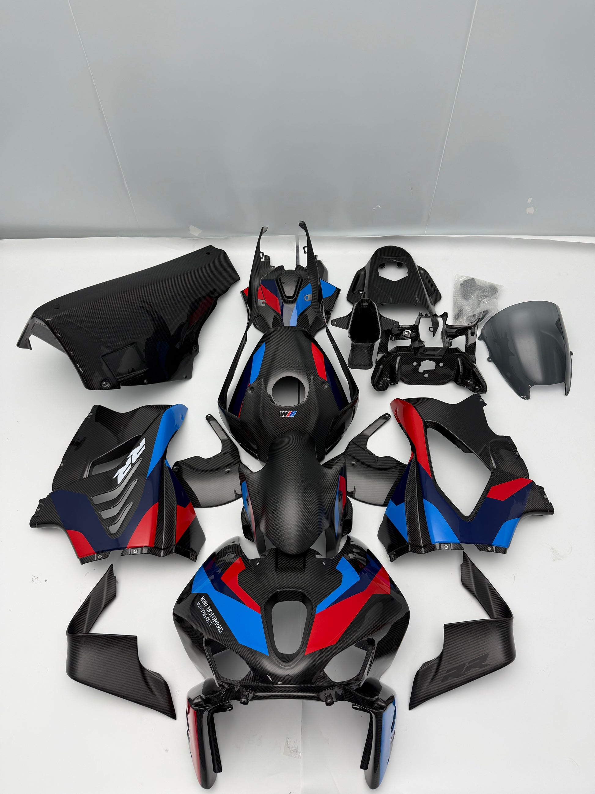 A set of motorcycle body panels and fairings laid out on a light beige fabric background. The parts are primarily black with a carbon-fiber texture, featuring the signature BMW M-style blue and red side stripes.
